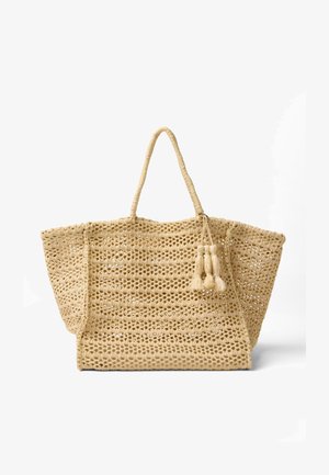 Beige gevlochten draagtas met opengewerkt patroon, twee handvatten en decoratieve kwastjes aan één zijde, afgebeeld op een witte achtergrond.