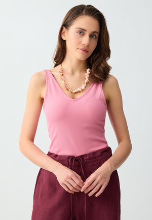 Femme aux cheveux bruns ondulés portant un haut rose sans manches, un pantalon taille haute bordeaux et un collier corail épais, mains détendues jointes.