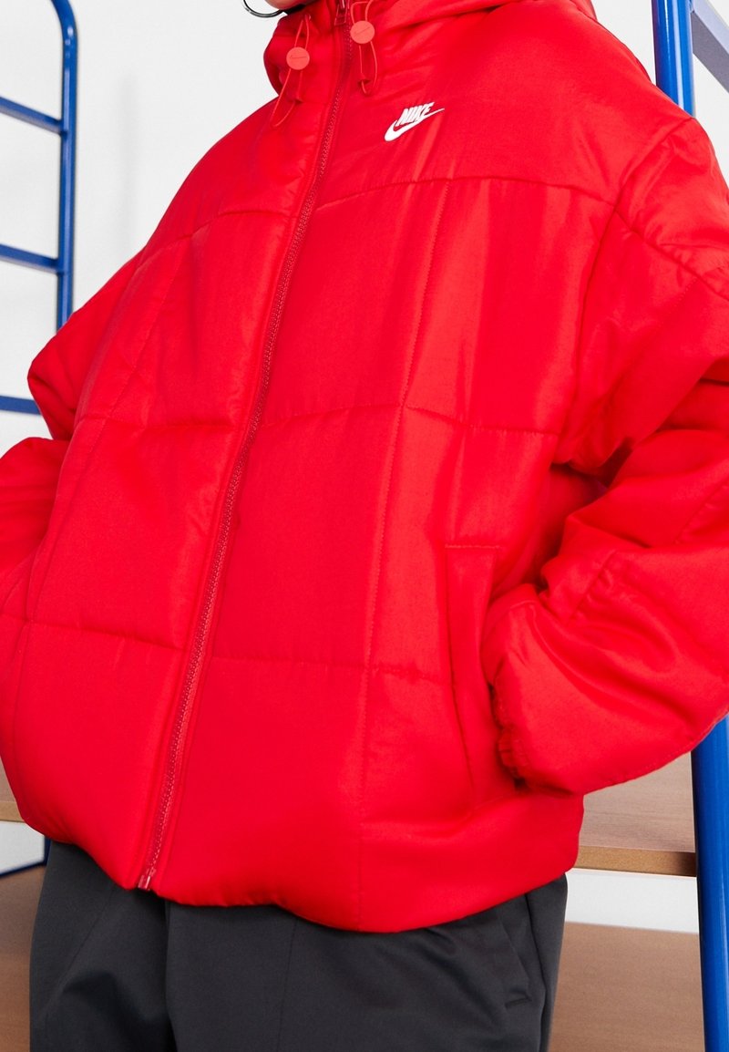 Röd vadderad jacka med främre dragkedja, hög krage och justerbar huva. Har kviltade sektioner och en liten vit logotyp på bröstet.