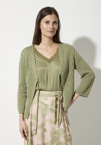 Lichtgroene gebreide cardigan met een open geweven ontwerp, lange mouwen en een ontspannen pasvorm, gedragen over een bijpassend satijnen top en bloemenshort.