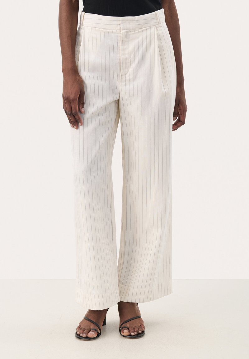 Pantalon à jambes larges crème avec fines rayures verticales, confectionné dans un tissu lisse. Présente des plis et une taille haute. Assorti à des sandales à talons noires.