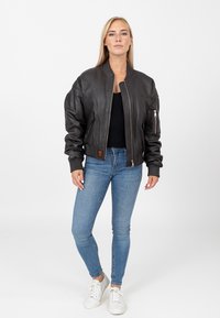 Brązowa skórzana kurtka bombowiec z prążkowanymi mankietami i pasem, z zamykanymi na zamek kieszeniami i pikowaną podszewką. Nosi się ją na czarnej bluzce.