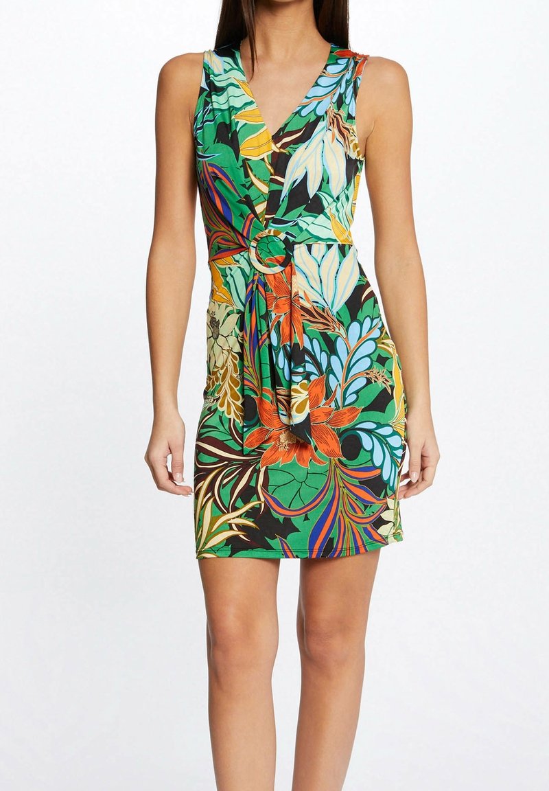 Ärmelloses, V-Ausschnitt Kleid mit tropischem Blumenmuster und einer Ringgürtel-Detail an der Taille, getragen von einer Frau, die vor einem schlichten Hintergrund steht.