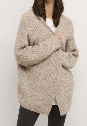 Kobieta w dużym beżowym dzianinowym swetrze narzuconym na biały top i czarne legginsy, trzyma sweter zamknięty rękami na wysokości klatki piersiowej.