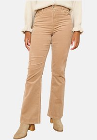 Ljusbruna kordflorsbyxor med utsvängda ben, slät yta, hög midja och framträdande sömnadsdetaljer, i kombination med beige klackboots som går upp till anklarna.