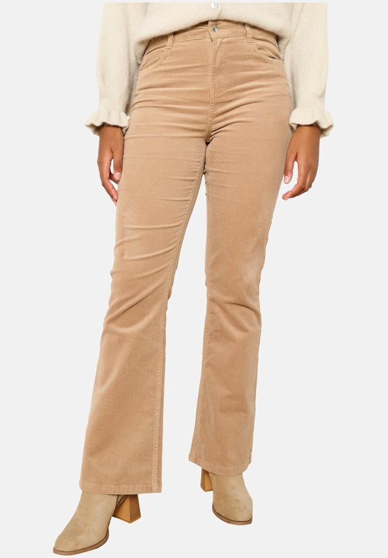 Ljusbruna kordflorsbyxor med utsvängda ben, slät yta, hög midja och framträdande sömnadsdetaljer, i kombination med beige klackboots som går upp till anklarna.