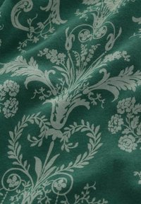 Zaļš audums ar baltām ziedu un viļņu dizainiem, ar sarežģītām detaļām, tostarp lapām un mazām ziediem uz strukturētas virsmas.