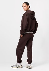 Hanorac maro supradimensionat și pantaloni de trening asortați dintr-un material moale, cu un detaliu de logo vizibil, asortați cu adidași gri și șosete albe.