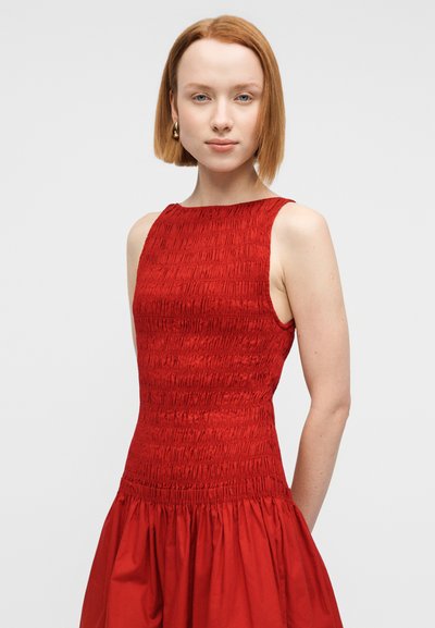 Κόκκινο φόρεμα με πτυχώσεις στο μπούστο και φαρδιά φούστα. Αμάνικο σχέδιο, υφασμάτινη υφή και μήκος μέχρι το γόνατο. Χρυσά σκουλαρίκια ως διακοσμητικά στοιχεία.