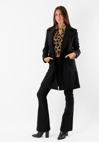 Cappotto lungo nero con revers e tasche, indossato sopra una maglietta con stampa leopardata. Pantaloni neri a campana e stivaletti con tacco completano l'outfit.