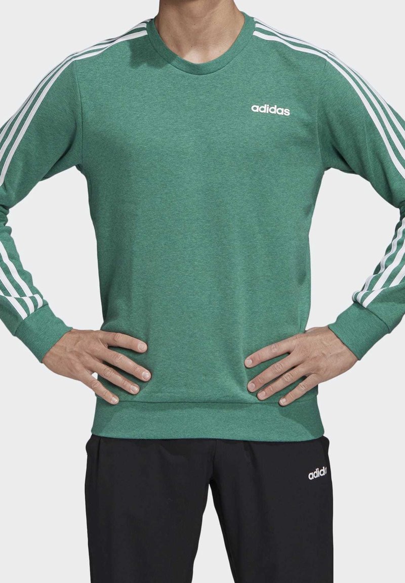 Homme portant un sweat-shirt vert Adidas avec des bandes blanches sur les manches et un pantalon Adidas noir, debout les mains sur les hanches.