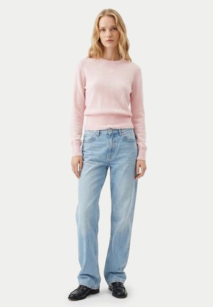 Blonde Frau steht frontal, trägt einen hellrosa Pullover, lockere hellblaue Jeans und schwarze Lederschuhe vor weißem Hintergrund.