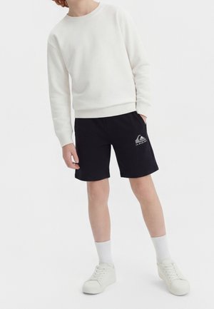 Osoba ubrana w białą bluzę z długim rękawem, czarne spodenki z logo, białe skarpetki i białe sneakersy, stojąca na tle jednolitego białego tła.