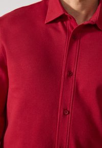 Camisa polo roja con cuello, cinco botones y una tela texturada. El diseño es sencillo, sin patrones ni acentos adicionales.