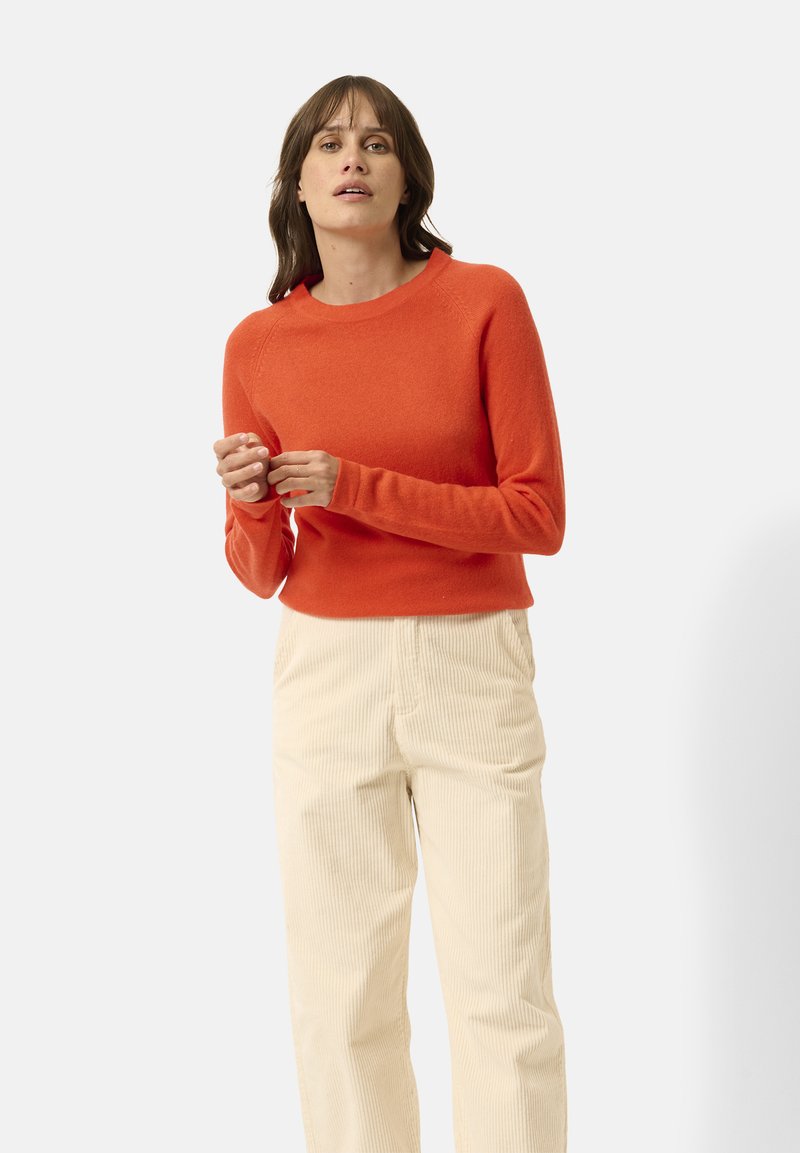 Pull en tricot orange avec des manches longues et un col ras du cou, associé à un pantalon en velours côtelé écru avec une rayure verticale texturée.