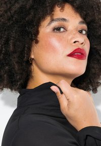 Schwarzes, hochkragen Kleidungsstück mit glatter Textur. Hat lockige Haare, kräftige rote Lippen und dezente Make-up Akzente. Die Hand greift den Kragenstoff.