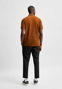 Brun polotröja med korta ärmar, ribbad krage, kombinerad med svarta byxor och grå skor, sedd bakifrån, mot en ljus bakgrund.