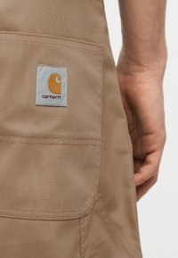 Τσέπη παντελονιού Carhartt σε χακί χρώμα με ραμμένη ταμπέλα λογότυπου, εμφανιζόμενη δίπλα στο χέρι και το μπράτσο ενός ατόμου πάνω σε απλό φόντο.
