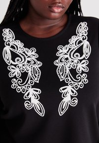 Black top featuring intricate white floral embroidery on both sides. Smooth texture with a round neckline. Detailed design elements are prominent.
