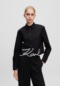 Zwarte overhemd met knoopsluiting en een puntig boord, voorzien van een witte geborduurde handtekening op de taille. Glad stof en getailleerd ontwerp.
