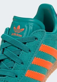 Turkos mockasinsneaker med orange zigzag-ränder, texturerade snören och en vadderad tunga med en präglad logo. Gummisula ingår.