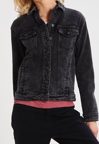 Svart denimjacka med knäppning, två bröstfickor och en texturerad yta. Har en klassisk krage och långa ärmar.