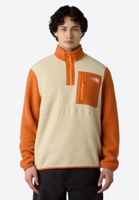 Fleece pullover met oranje en crèmekleurige panelen, half-rits kraag, rits borstzak en het logo van The North Face op de zak. Zachte textuur.