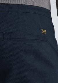 Pantalons en velours côtelé bleu marine avec une poche arrière dotée de détails de couture et d'une petite étiquette "PINE LEGEND" en or. Tissu texturé.