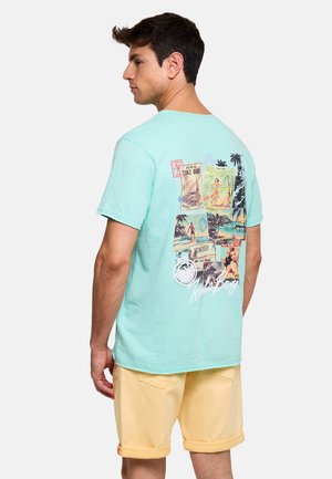 Hombre con camiseta turquesa claro con estampados de postales vintage de Hawái y pantalones cortos beige, de pie frente a un fondo blanco.