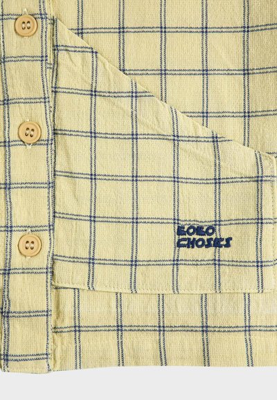 Tissu jaune clair avec un motif en grille bleue, trois boutons beige, et une poche pliée brodée de "SOLO CHOICES" en fil bleu.