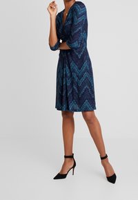 Polvipituinen, kietaisutyylinen mekko tummansinisenä, jossa on petrolinsininen siksak-kuvio. Valmistettu teksturoidusta kankaasta; siinä on kolme neljänneksen hihaa ja liehukehelma.