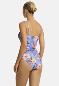 Una donna con un'acconciatura a bun indossa un costume da bagno blu floreale, in mostra un motivo tropicale. È rivolta di spalle rispetto allo spettatore.