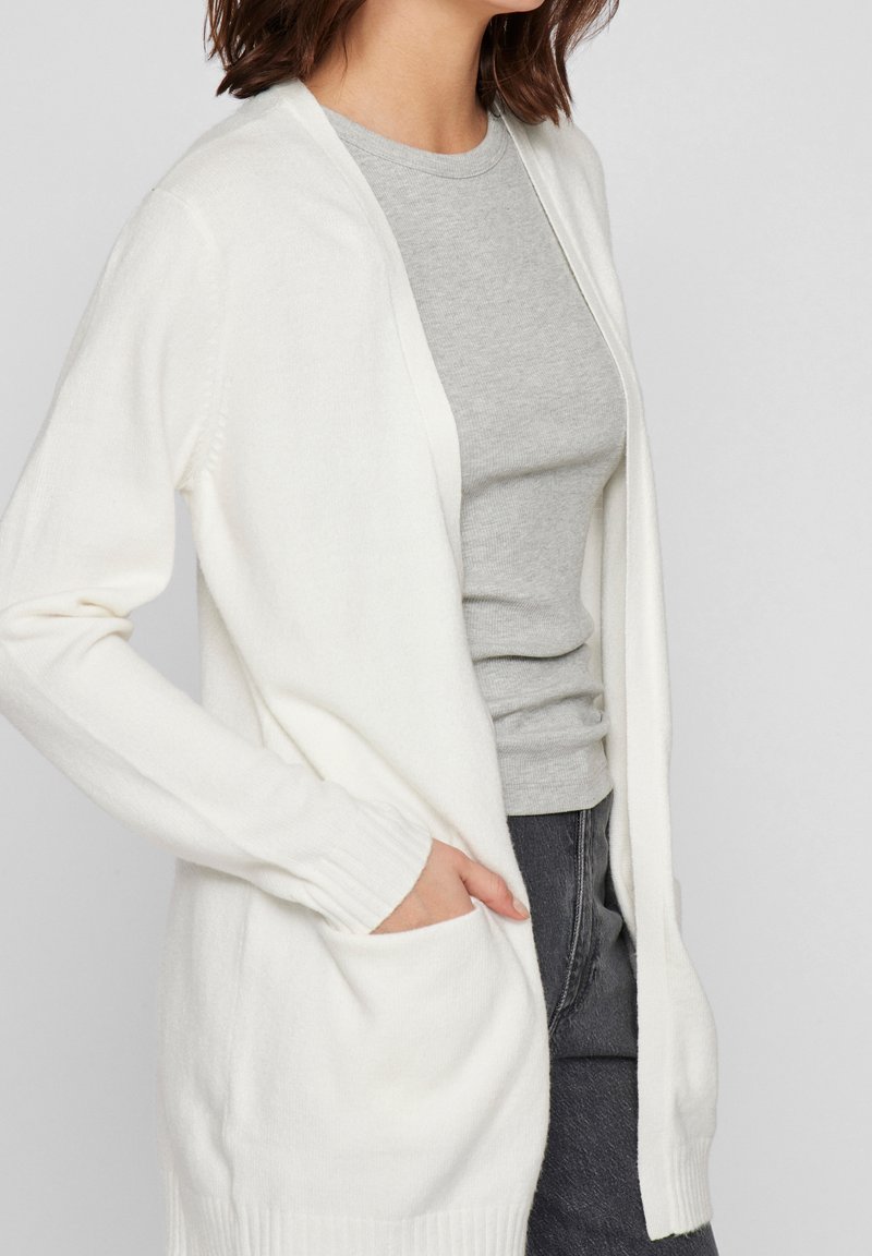 Femme portant un cardigan blanc ouvert à l'avant avec des poches, par-dessus un haut gris à manches courtes côtelé, et un jean foncé, la main dans la poche du cardigan.