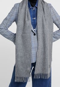 Szary wełniany szalik z frędzlami, o miękkiej fakturze. Ułożony na ramionach osoby, w zestawieniu z marynarką w kratę.
