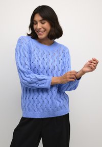 Pulover tricotat de culoare albastru deschis, cu un model în valuri și țesătură deschisă. Guler tip crew și manșete ribbate. Asortat cu pantaloni negri.