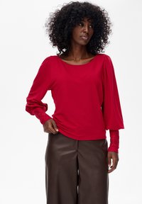 Kobieta z kręconymi włosami, ubrana w czerwoną bluzkę z długim rękawem i bufkami oraz brązowe skórzane spodnie, patrzy w lewo.