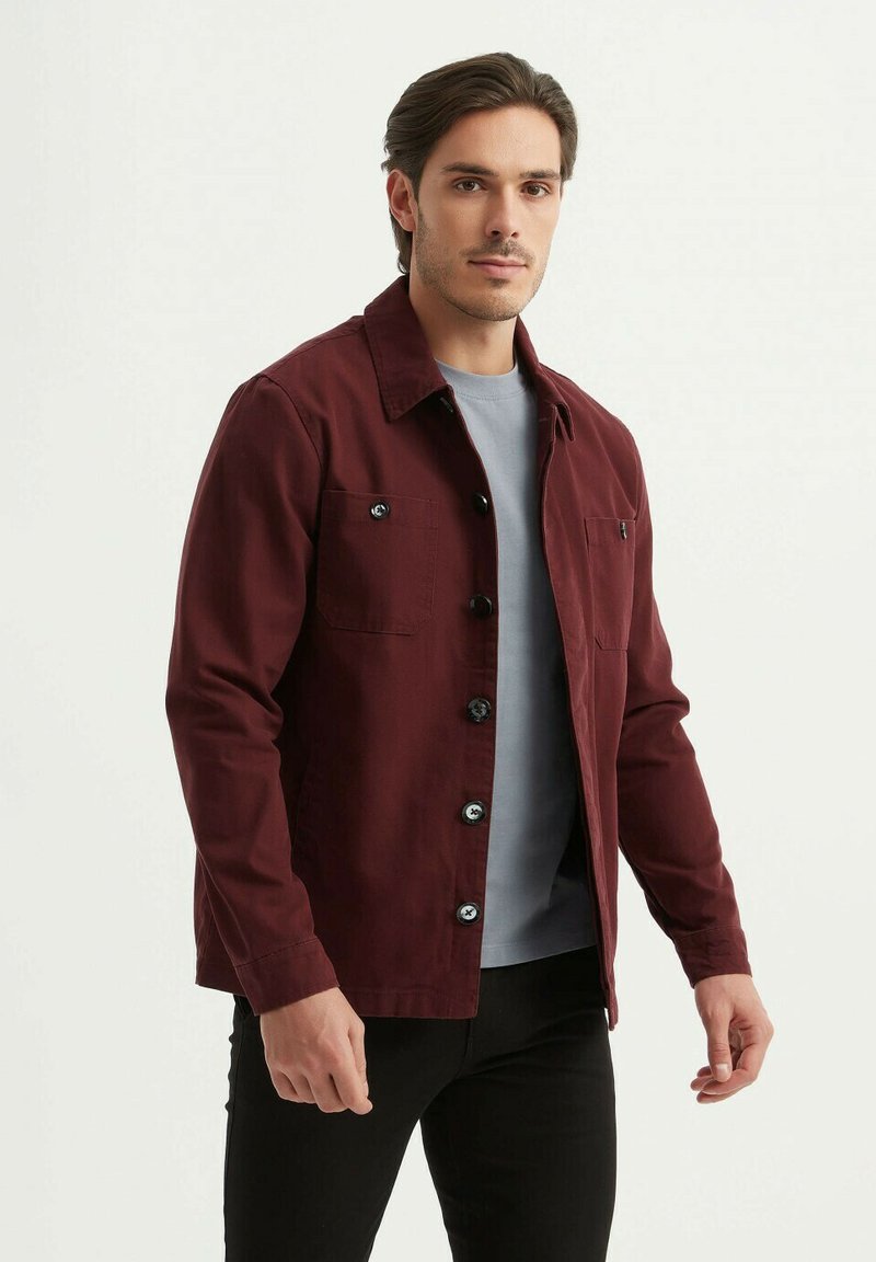 Veste bourgogne en coton, avec un devant boutonné, deux poches poitrine, et un col, portée sur une chemise grise.