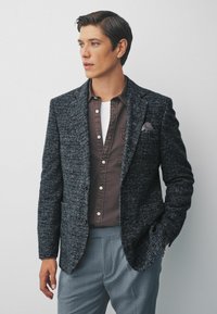 Sacou texturat, gri închis, cu rever tăiat, purtat peste o cămașă cu nasturi maro. Pantaloni gri completează ținuta. Un batiste notabil în buzunar.