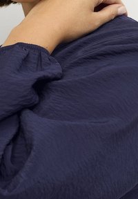 Tissu bleu marine avec une texture froissée ; détail de poignet froncé ; avant-bras et main partiellement révélés touchant le tissu.