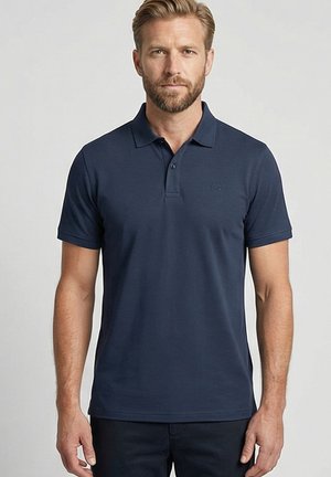 Man met korte baard die een effen donkerblauw poloshirt met korte mouwen draagt, staand tegen een effen lichte achtergrond.