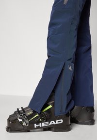 Pantaloni de schi navy cu fermoare laterale purtați peste bocancii de schi HEAD negri și verzi pe un fundal alb.