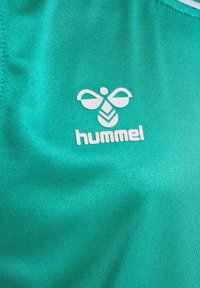 Camisa deportiva turquesa hecha de tela suave, con un logotipo blanco de "hummel" y un diseño de abeja estilizado que transmite una estética moderna.