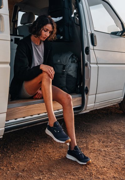 Černé sportovní boty se šedými akcenty a gumovými podrážkami, které má na sobě osoba sedící na dřevěném schodu dodávky. Nedaleko je šedý batoh.