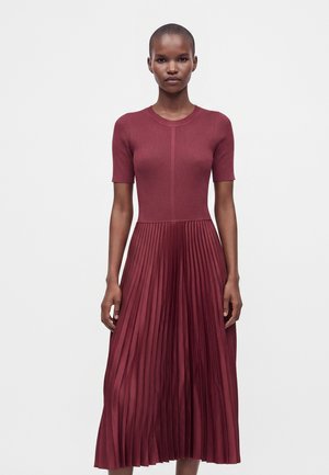 Femme aux cheveux courts portant une robe bordeaux avec un haut côtelé ajusté et une jupe plissée, debout devant un fond clair uni.