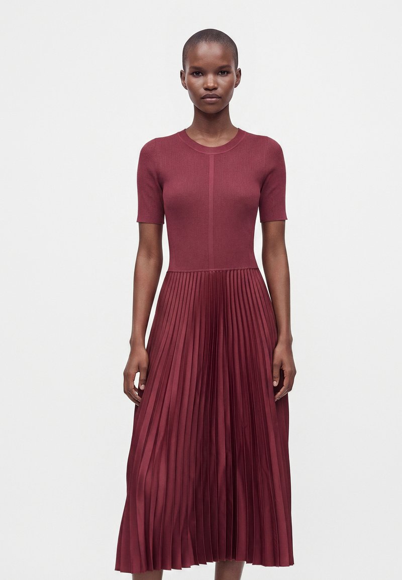 Femme aux cheveux courts portant une robe bordeaux avec un haut côtelé ajusté et une jupe plissée, debout devant un fond clair uni.