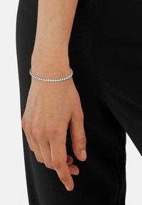 Brazalete de plata con gemas circulares transparentes dispuestas en una línea continua, llevado en una mano sobre un fondo de tela negra.