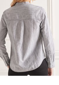 Camicetta a righe grigie e bianche con orlo arrotondato, polsini con bottoni e yoke posteriore. Indossata con pantaloni neri. Il tessuto appare liscio.