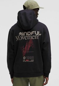 Mann mit schwarzem Hoodie, auf dem "Mindful Movement" und ein rohes Wellenmotiv abgebildet sind, beigefarbener Kappe und grünen Hosen, von hinten vor einem schlichten Hintergrund.