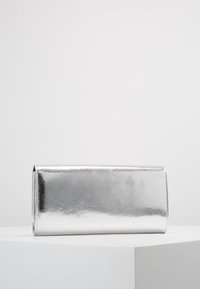 Silverfärgad, metallisk rektangulär clutchväska som vilar på en vit yta mot en enfärgad ljusgrå bakgrund.