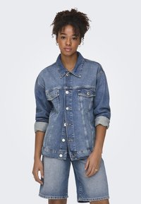 Chaqueta de mezclilla en azul claro con cierre de botones, dos bolsillos en el pecho y mangas enrolladas. Combinada con unos pantalones cortos de mezclilla a juego.